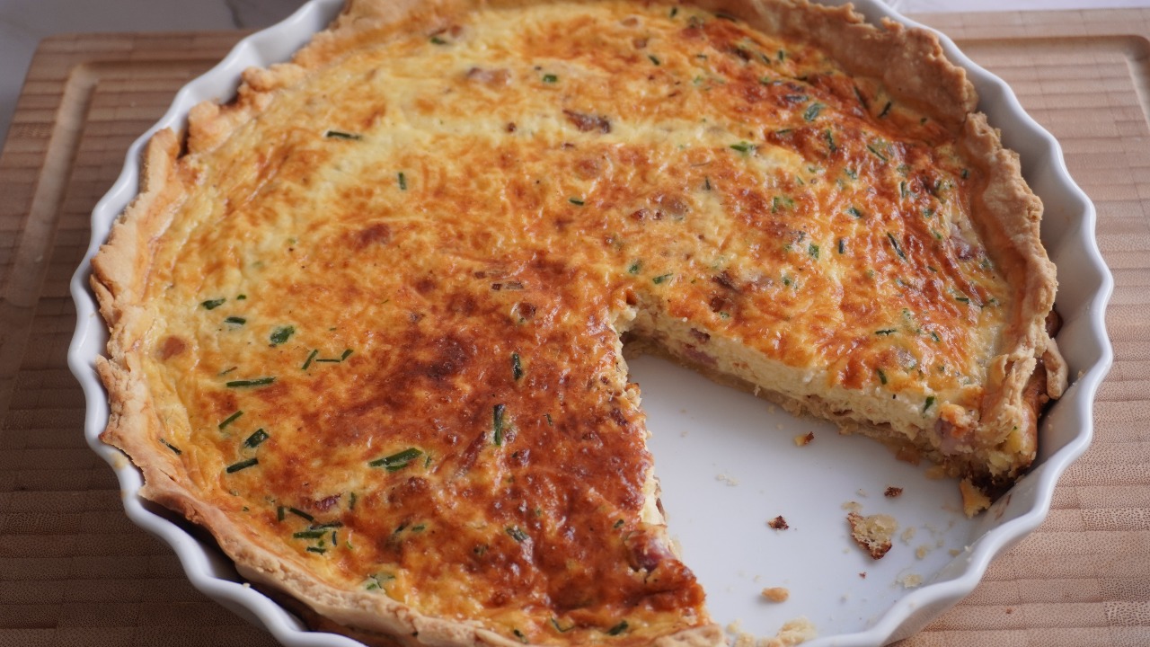 Homemade quiche lorraine with golden Gruyère top and chives in a white fluted tart dish, one slice removed showing the bacon and onion filling and crisp butter crust.
